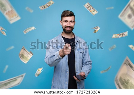 Young cheerful man in black t-shirt and denim jacket with phone in his hand. Guy browsing internet on smartphone and expresses happiness. Blue background. Money rain. Falling hundred dollar banknotes.