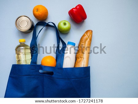 A set of food products which consists of milk, red pepper, a can of canned food, apple, orange, sunflower oil and a baguette in a blue bag, which lies on a background of marine color.View from above.