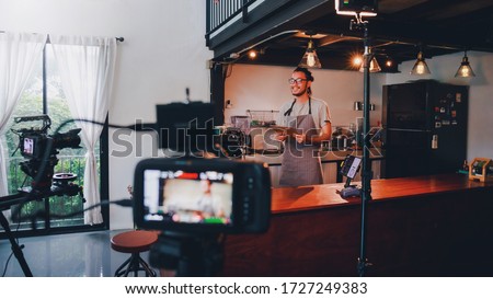 Asian barista man standing in front of the camera and recording vlog video live streaming at bar counter in coffee shop interior background.Sale and promotion online marketing business concept.