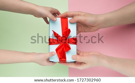 Top view of children's and women's hands holding a white gift box with a red ribbon on a pink and green background. Flat lay. Gift for birthday, Christmas, Valentine's day, New year. 