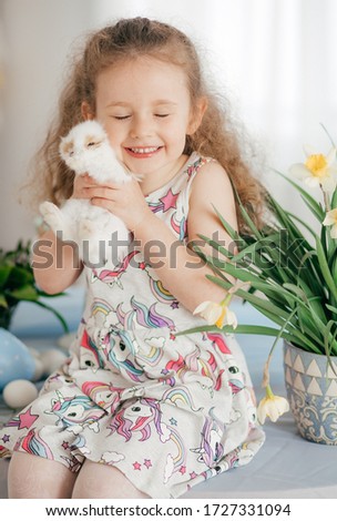 Stock Photo -  Little girl play with rabbit indoors in spring. Decorated home and spring flowers. Family celebrating Easter