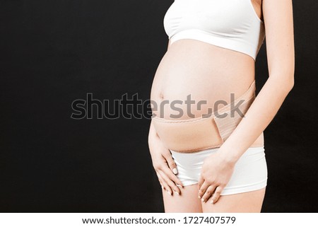 Close up of pregnant woman in underwear with supporting bandage against backache at black background with copy space. Orthopedic abdominal support belt concept.