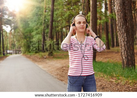Horizontal photo of cheerful delighted good looking young female looking ahead, having headphones, listening to music, wearing jeans and sweatshirt, spending free time in peace. Rest concept.