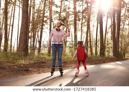 Outside picture of cheerful energetic young female rollerskating with her little daughter, enjoying nature, being at forest, having headphones around neck, leading active lifestyle. Hobby concept.