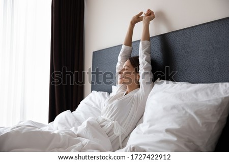 Single rested renewed after deep healthy night sleeping 30s woman wake up do stretching exercises seated in bed, satisfied female raised hands closed eyes enjoy early morning at home hotel room alone Royalty-Free Stock Photo #1727422912