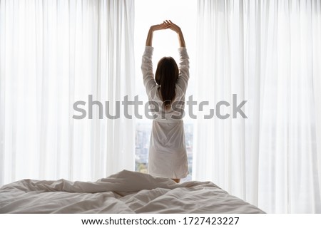 Rear view woman in white night robe woke up stands near window looks out the window enjoy city view, raised hands doing morning exercises stretches body muscles, starting new day, well-being concept Royalty-Free Stock Photo #1727423227