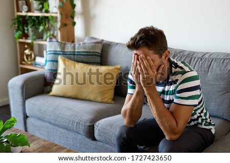 Young man looking upset while sitting alone on his living room sofa at home with his face in hands Royalty-Free Stock Photo #1727423650