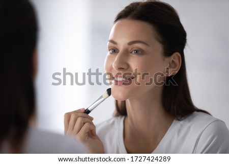 Close up beautiful young woman reflected in mirror smile applying concealer foundation cream using natural bristle make-up professional brush, facial makeup, cosmetics tool, beauty, skincare concept Royalty-Free Stock Photo #1727424289