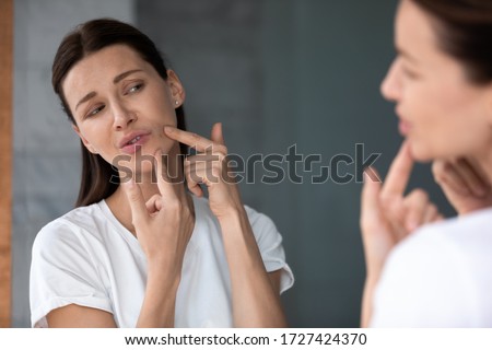 Over shoulder view upset female face reflected in mirror, woman touch face squeezes pimple caused by hormonal imbalance changes, stress or diet. Skin problem, jawline acne or new mole appears concept Royalty-Free Stock Photo #1727424370