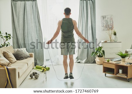 Back view of young man in activewear jumping with skipping-rope while standing in front of window of living-room during workout Royalty-Free Stock Photo #1727460769
