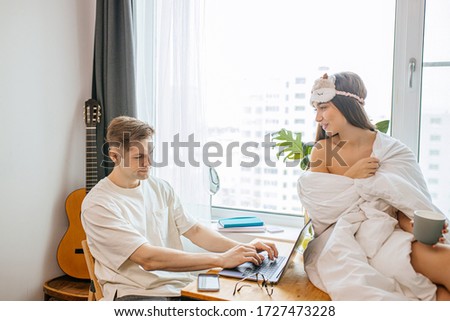 young caucasian woman support husband during his freelance work at home, female wrapped only in blanket, man sit with laptop. in the morning