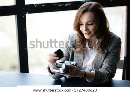 Attractive woman in a plaid jacket holds a manual lens in her hand, unscrewed from the old retro camera, sitting at the table.