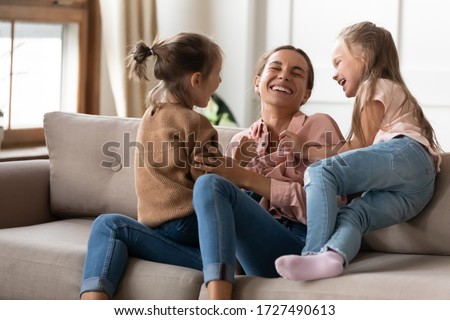 Happy mother playing with two little daughters, sitting on couch at home, cute preschool girls tickling laughing mum, family having fun, spending weekend together, playing funny game Royalty-Free Stock Photo #1727490613
