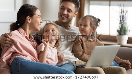 Happy parents with two little daughters using laptop at home, hugging and talking, watching cartoons, shopping online, making video call, family having fun with computer together