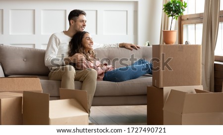 Happy young couple relaxing on couch on moving day in living room with cardboard boxes, dreamy wife and husband looking in window, dreaming about good future, excited by relocation into new house Royalty-Free Stock Photo #1727490721