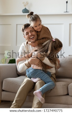 Funny little girls hugging cuddling smiling laughing father sitting on comfortable couch in living room, excited father having fun with two adorable little daughters, enjoying free time together Royalty-Free Stock Photo #1727490775