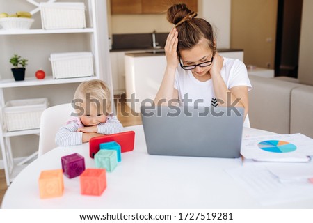 A young mother is tired of working on a laptop, her little daughter is sitting next to her and watching favorite cartoon on the phone. Work at home