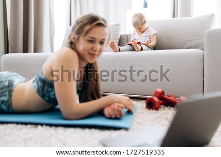 A small cute child sitting on a couch is watching a cartoon on the phone, and mom in the foreground, in defocus, does sports and watching in a laptop