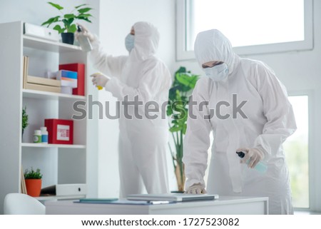 Protective measures. Medical worker in protective clothing and medical mask disinfecting table, another worker standing behind, cleaning closet Royalty-Free Stock Photo #1727523082