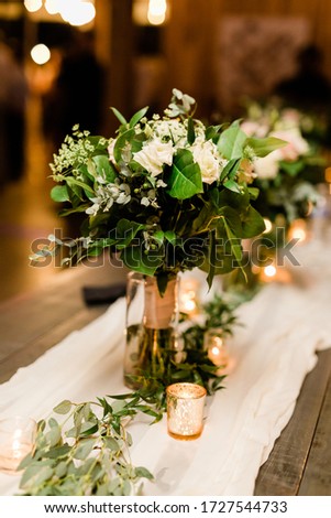 Bridal Bouquet with White Roses, Baby's Breath and Eucalyptus Leaves in a Vase at the Head Table at a Wedding with Votive Candles and a White Cheesecloth on a Wood Table Royalty-Free Stock Photo #1727544733