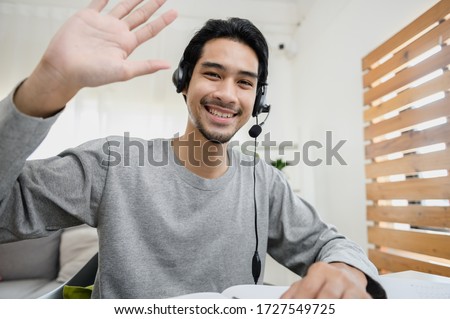 Portrait photo of smart Asian guy using video call communicate with business colleague friend from home during self isolation from coronavirus outbreak crisis. Asian man waving hand looking at camera. Royalty-Free Stock Photo #1727549725