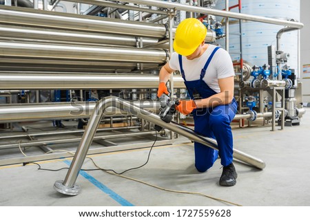 Factory worker in yellow hard hat operate with stainless steel pipe sander polishing machines. Remove spot welds or eliminate deep scratches. Grind and polish up to high-gloss finish. Indoor. Royalty-Free Stock Photo #1727559628