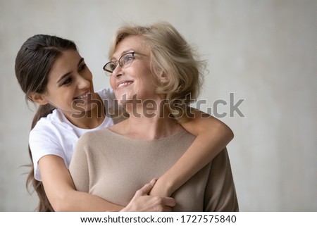 Close up headshot portrait picture of adult daughter hugs from behind happy mature mother enjoying tender moment looking at each other. Smiling woman and older mum embracing having fun together.