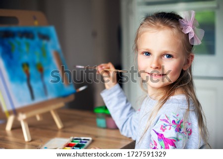 Beautiful little girl draws paints on paper at home sitting at the table. Training in drawing. Creative quarantined activities. Hobbies for children