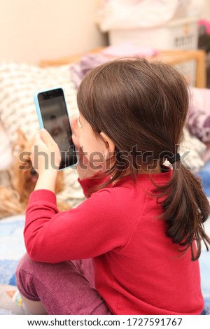 A little white girl with a ponytail takes a picture of her sister on a cell phone against the background of a regular apartment