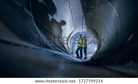 Two Heavy Industry Engineers Walking Inside Pipe, Use Laptop, Have Discussion, Checking Design. Construction of the Oil, Natural Gas and Biofuels Transport Pipeline. Industrial Manufacturing Factory Royalty-Free Stock Photo #1727599216