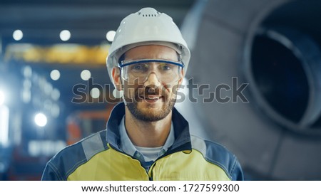 Portrait of Smiling Professional Heavy Industry Engineer / Worker Wearing Safety Uniform, Goggles and Hard Hat. In the Background Unfocused Large Industrial Factory Royalty-Free Stock Photo #1727599300