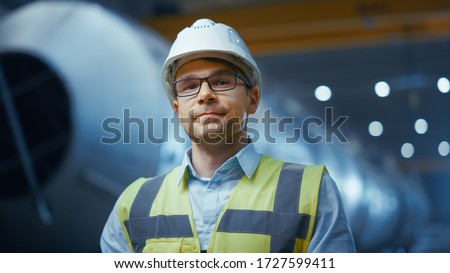 Portrait of Young Professional Heavy Industry Engineer / Worker Wearing Safety Vest and Hardhat Smiling on Camera. In the Background Unfocused Large Industrial Factory where Welding Sparks Flying. Royalty-Free Stock Photo #1727599411