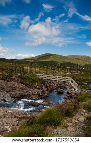 Beautiful green mountains in Scotland in the Highland region. A river flows between the mountains and a footbridge crosses the river. Blue and cloudy sky.