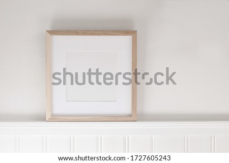 Closeup of square empty wooden picture frame on shelf. White beadboard wainscot wall paneling background. Scandinavian interior, home design. Art concept. Artistic mockup scene. Front view.