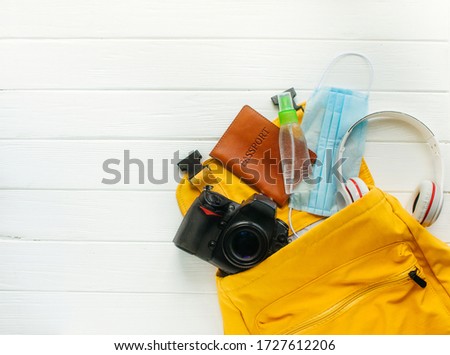 Travel backpack with face mask, sanitizer, passport, photocamera and headphones on white wooden background with blank space for text. Packing for a travel after the end of pandemic COVID-1.