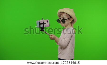 Girl child makes selfie on mobile phone using selfie stick. Kid with pink hair in hat and sunglasses using smartphone social network app for video call, blogging. Green screen at studio. Chroma key