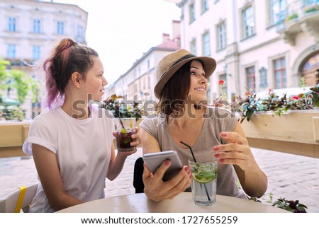 Talking and drinking soft drinks mother daughter teenager, smiling women sitting in outdoor cafe, with smartphone, looking away. Friendship and communication of parent and child of teenager