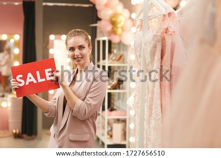 Waist up portrait of beautiful young woman holding SALE sign while posing in clothing boutique, copy space