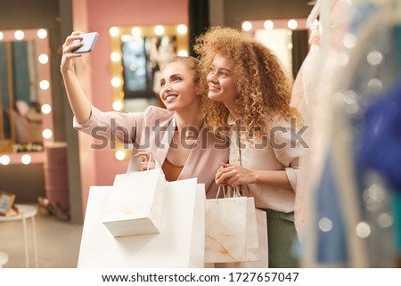 Waist up portrait of two young women taking selfie via smartphone while enjoying shopping in clothing store, copy space