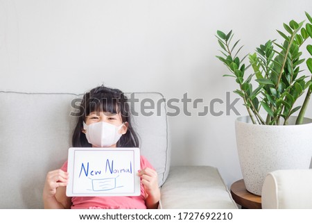 Kid girl holding tablet and writing NEW NORMAL word with face mask picture showing at home.New normal Post covid-19 crisis change human behavior.Concept for Stay home,Wellness health, Digital takeover Royalty-Free Stock Photo #1727692210