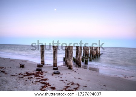 Dilapidated pier leading into the ocean on the beach of Port Royal in Naples, Florida at sunrise as the full moon sets. 