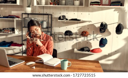 Portrait of young female worker receiving orders online or via telephone while sitting in the office. Woman working at custom T-shirt, clothing printing company. Horizontal shot. Selective focus