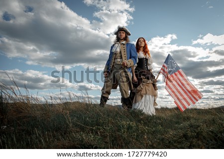 Man in form of officer of War of Independence and girl in historical dress of 18th century. July 4 is US Independence Day. Couple of patriots freedom fighters in outdoor on background cloudy sky Royalty-Free Stock Photo #1727779420
