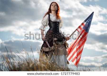 Girl in historical dress of 18th century with flag of United States. July 4 is US Independence Day. Woman of patriot freedom fighter in outdoor on background cloudy sky Royalty-Free Stock Photo #1727779483