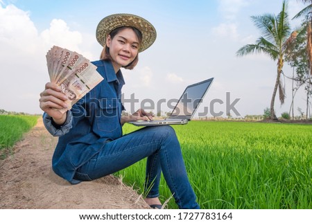 Happy asian farmer woman sitting and using laptop computer with holding banknote money at green rice farm