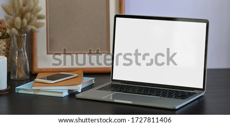 Photo of white blank screen computer laptop putting on black working desk and surrounded by stack of notebook, smartphone, empty picture frame, wild grass in vase and coffee cup.