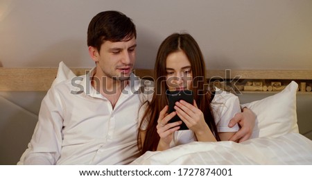 a young couple is sitting in bed with white linens. the woman looks intently at her smartphone. the guy comments. they smile