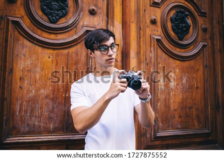Young male photographer amateur in spectacles making pictures on town during free time, skilled hipster guy traveler in casual wear taking photos of historical places of city holding camera