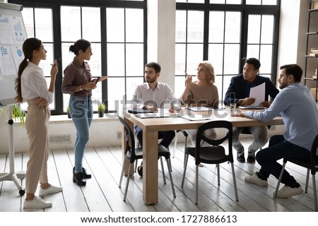 Indian and caucasian businesswomen flip charts presentation new project in boardroom at company meeting. Confident auditors speaks with mature and black partners about business using board and graphs. Royalty-Free Stock Photo #1727886613