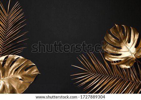 Shiny glossy golden painted tropical date palm and monstera leaves creatively arranged on black paper background. Empty space for copy, room for text. Trendy luxury border frame flatlay design.